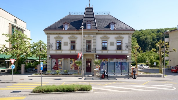 Un bâtiment à deux étages de style architectural classique, caractérisé par une façade symétrique, une entrée centrale avec une porte rouge et un petit balcon au-dessus de l’entrée.