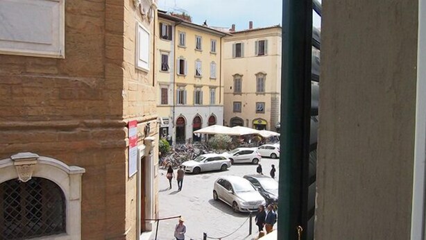Un patio con coches aparcados, algunas personas caminando y edificios con ventanas de arco y balcones.