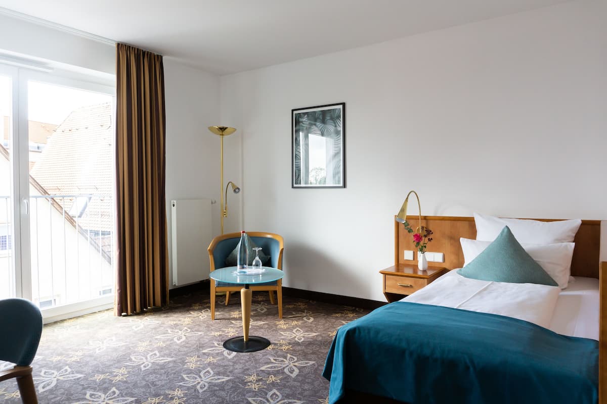 Ein Hotelzimmer mit Bett, Sessel, Tisch, Stehlampe und einem Fenster mit Vorhängen.
