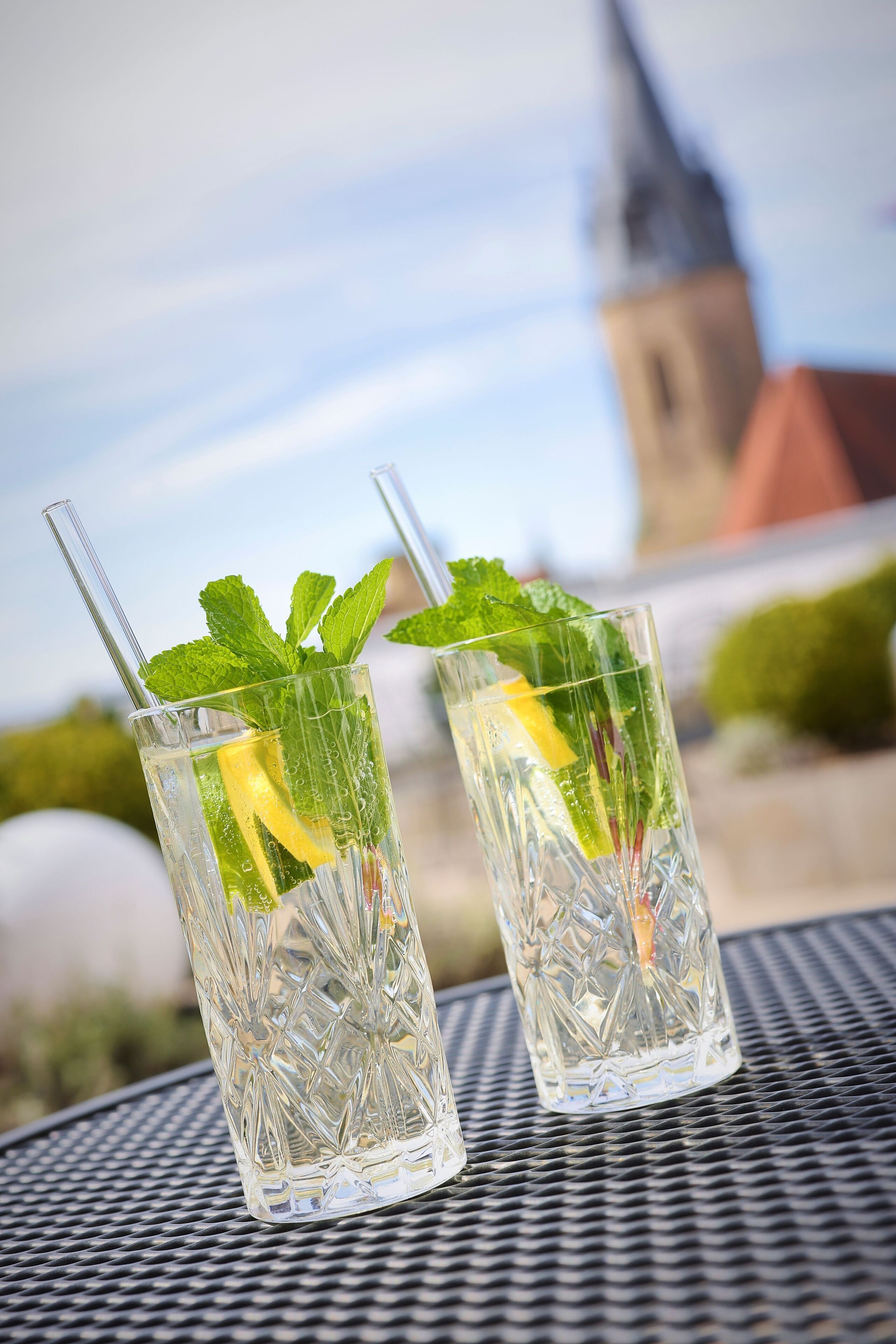 Two clear glasses with mint leaves and a lemon slice on a metal table with a church spire in the background.