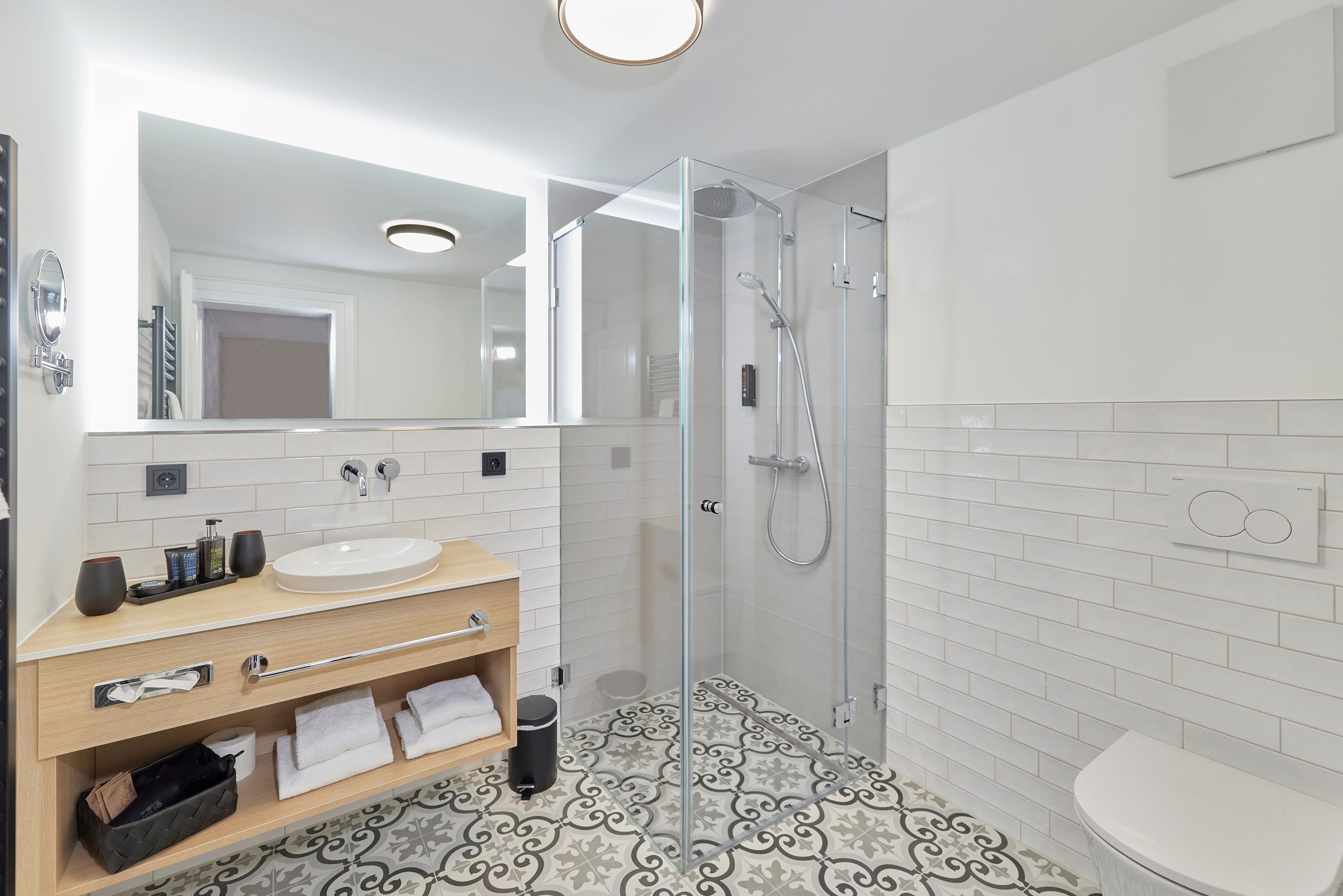 Un baño moderno con ducha de vidrio, tocador con lavamanos y espejo, y baldosas de suelo con diseño.