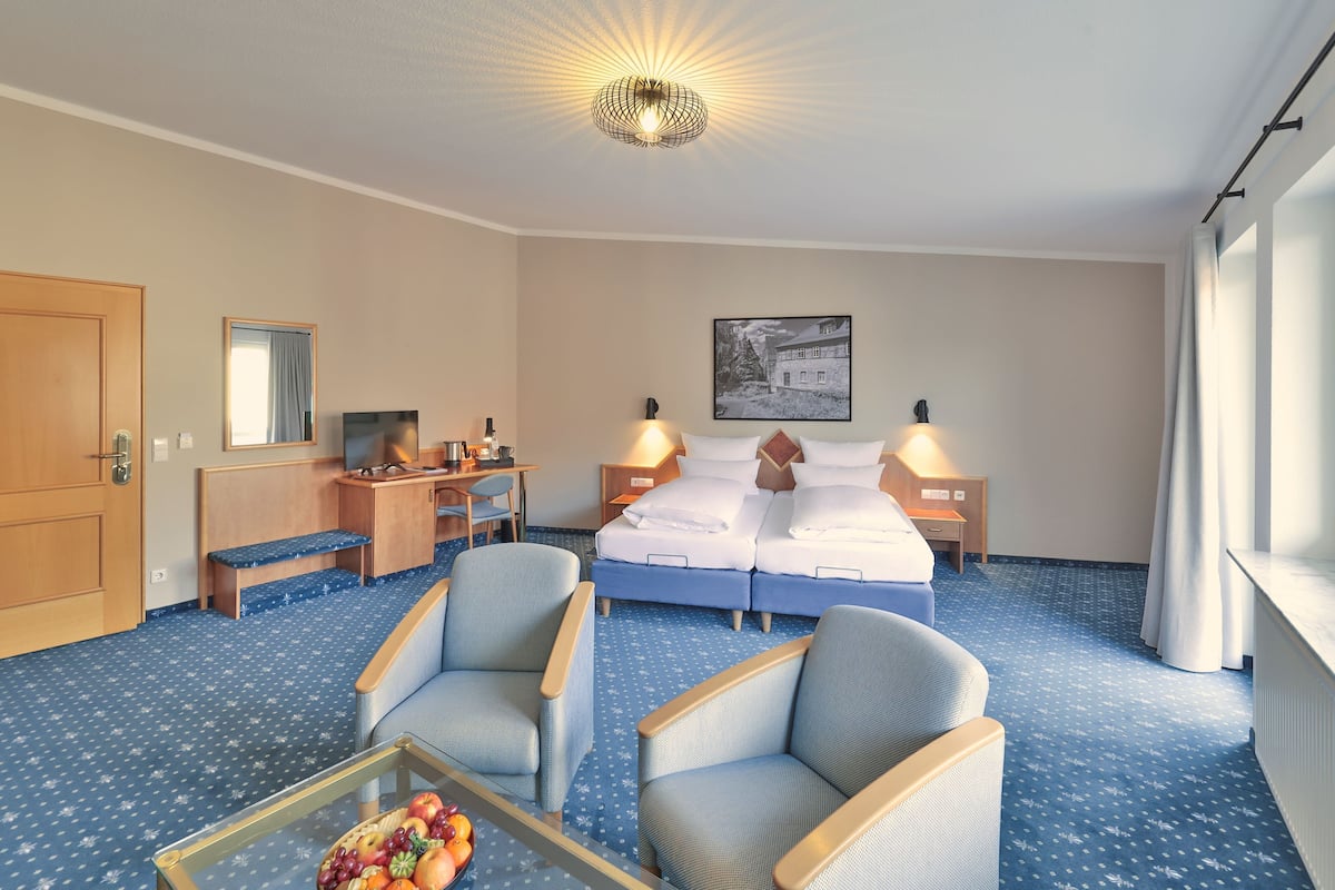 Ein Hotelzimmer mit einem Bett, zwei Nachttischen, einem Schreibtisch mit Stuhl, einer kleinen Sitzecke mit zwei Sesseln, einem Glastisch mit Früchten und einem großen Fenster mit Vorhängen.