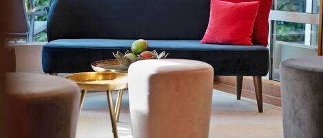 Una sala moderna con un sofá azul oscuro, una almohada roja y una bandeja dorada con frutas. Hay dos otomanas, una blanca y una gris, y un ventanal que ofrece vistas a la vegetación exterior.