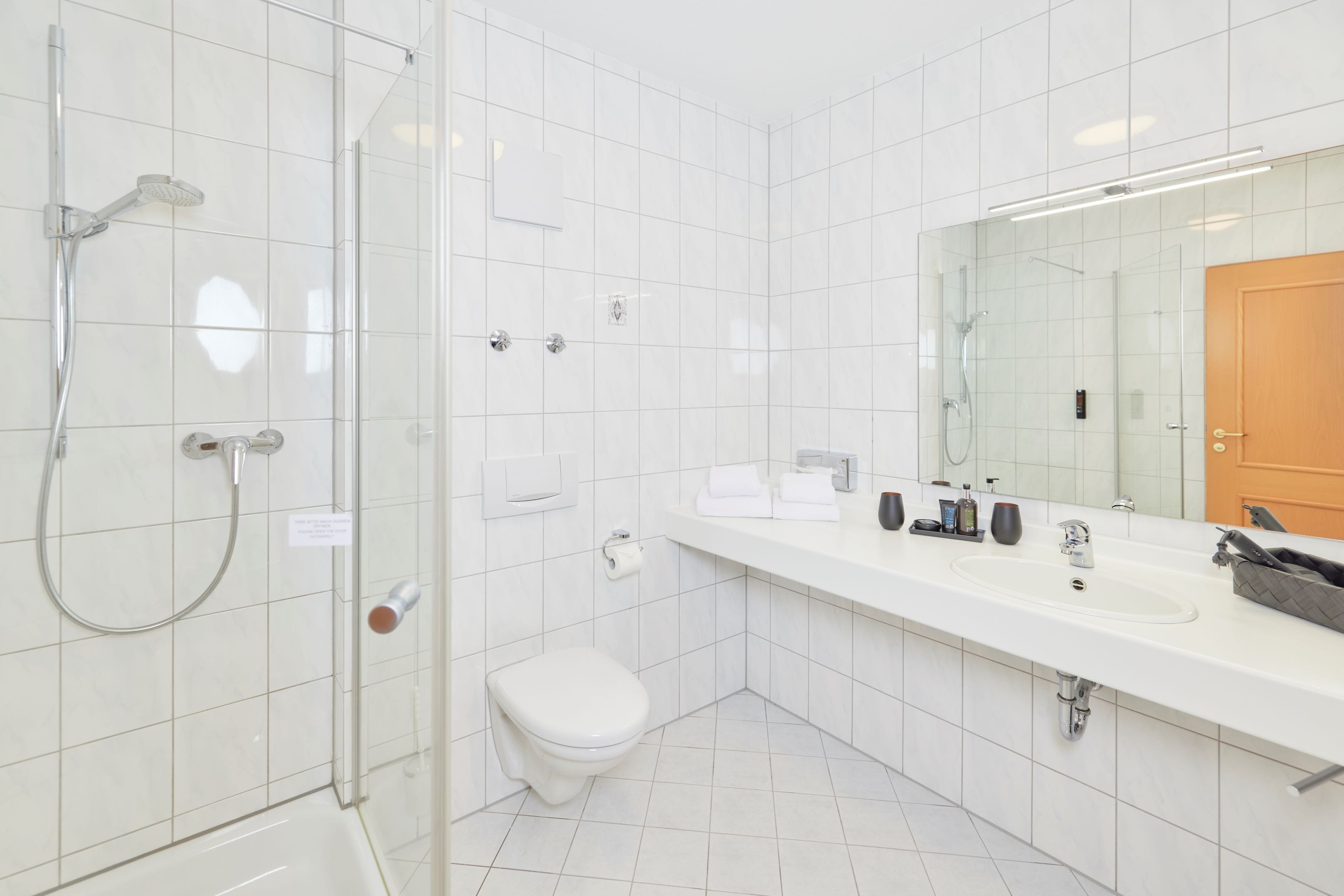 A modern bathroom with a glass-enclosed shower, a white toilet, and a large mirror above a white sink with various toiletries.
