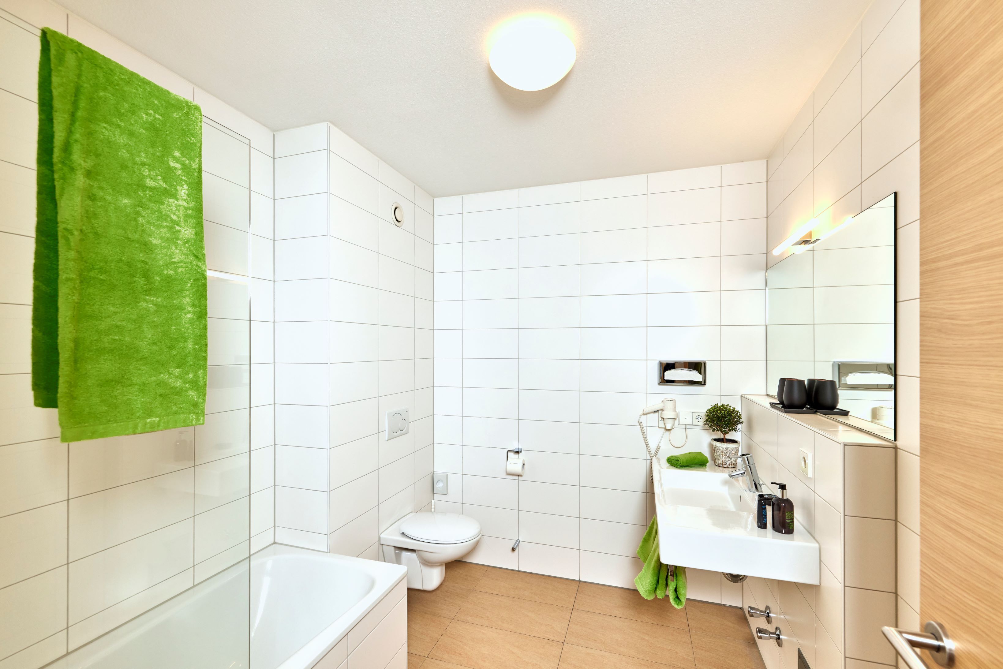 A modern bathroom with white tiles, a green towel, a toilet, a sink, and a mirror.