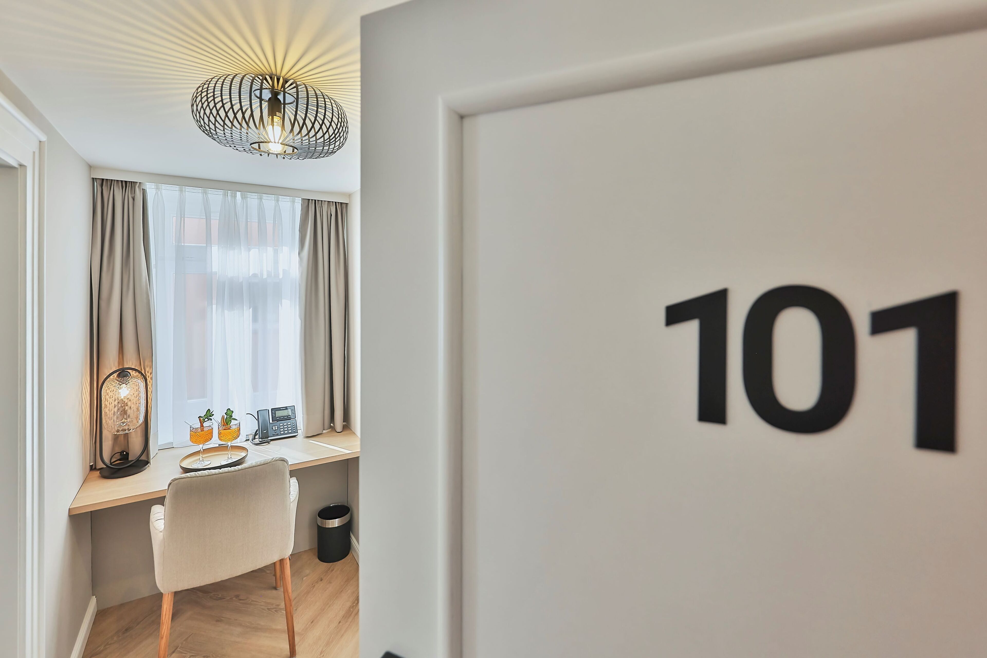 A hotel room with a desk, a chair, a lamp, a phone, and a trash bin. The room has a large window with curtains and a ceiling light fixture. The room number is 101.