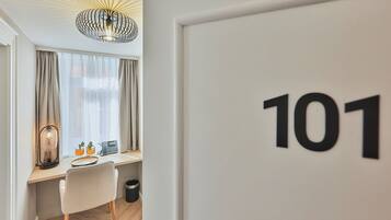 A hotel room with a desk, a chair, a lamp, a phone, and a trash bin. The room has a large window with curtains and a ceiling light fixture. The room number is 101.