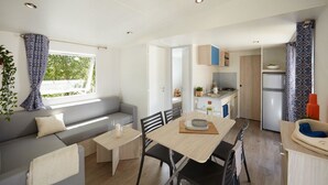 A modern, open-plan living and dining area with a sofa, dining table, chairs, and a kitchen in the background. - Camping Victoria (Canet de Mar)