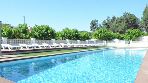 A rectangular swimming pool with clear blue water, surrounded by white lounge chairs and a white fence. - Camping Victoria (Canet de Mar)
