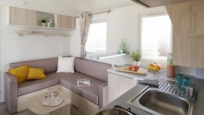 A compact living space with a kitchenette, a sofa with cushions, a small coffee table, and a window with curtains. - Camping Victoria (Canet de Mar)