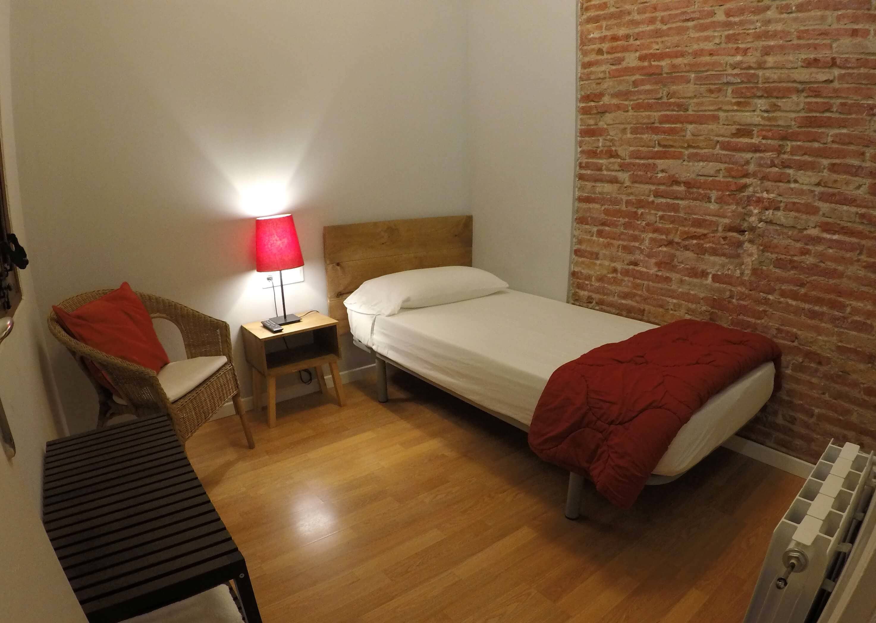 A small, cozy room with a single bed, a nightstand with a lamp, a wicker chair, and a radiator.