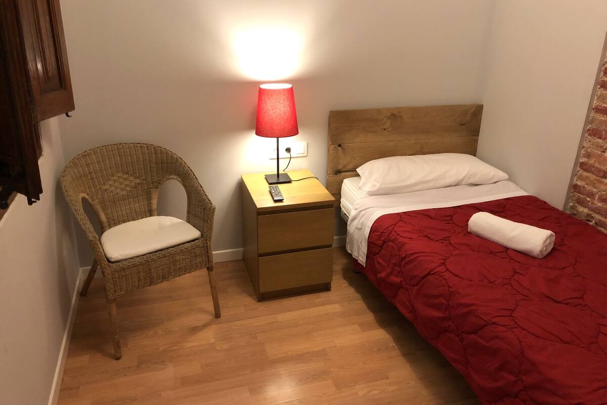 A small, cozy room with a wooden floor, a wicker chair, a nightstand with a red lamp, and a bed with a red blanket and white pillow.