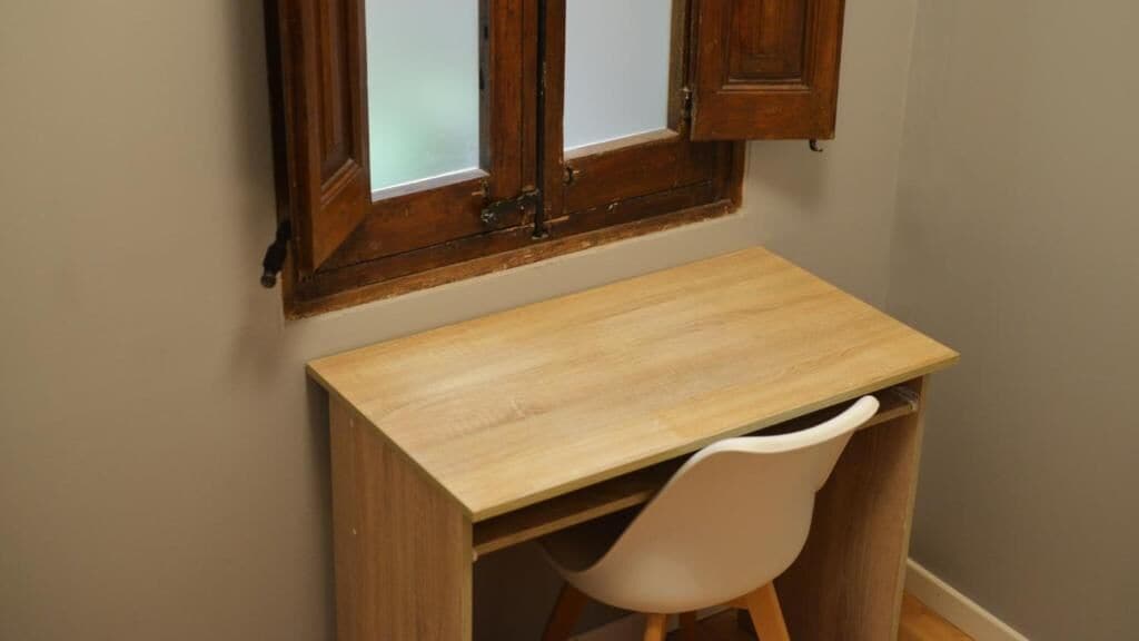 Un bureau en bois avec une chaise blanche, dans une pièce aux murs de couleur beige et dotée d’une fenêtre avec des volets en bois.