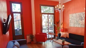 A living room with a red wall, a black sofa, a red armchair, a wooden coffee table, and a chandelier. - Hostal Paraiso (Barcelona)