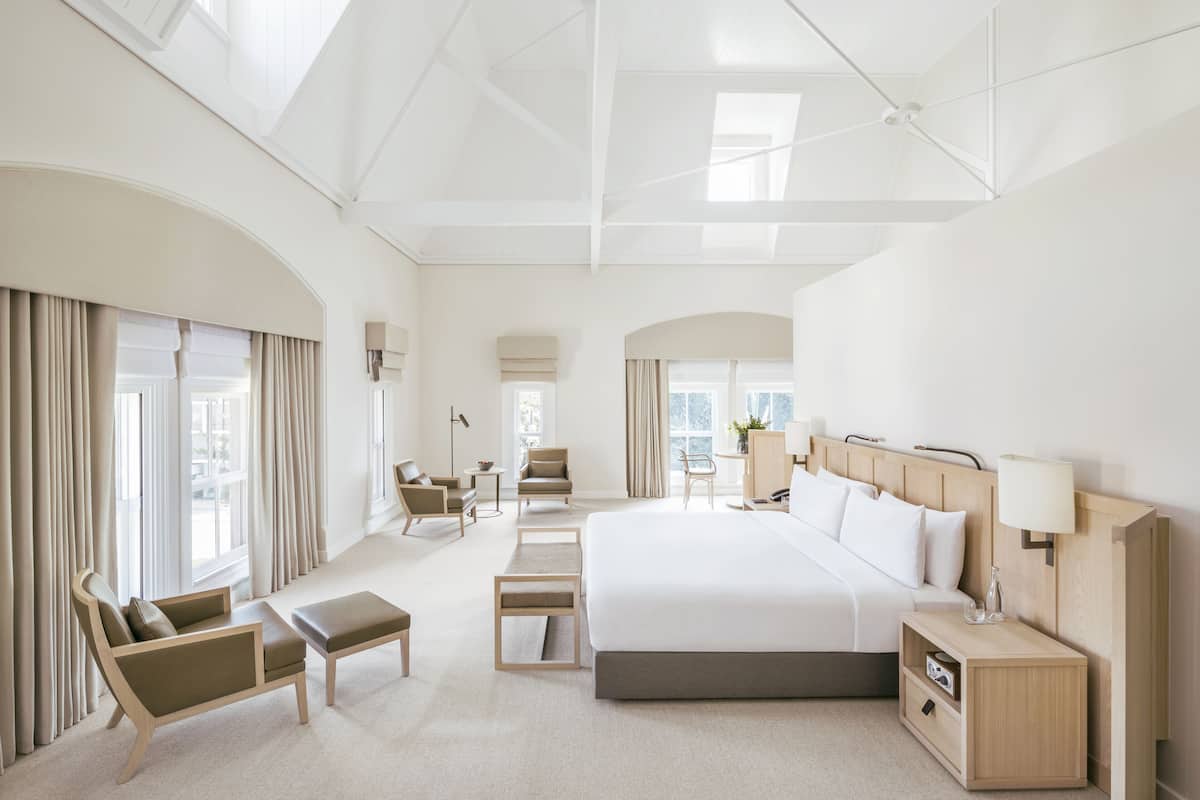 Ein modernes Hotelzimmer mit einem großen Bett, einer Sitzecke mit einer Couch und Sesseln, einem kleinen Schreibtisch und Blick auf die Stadt durch große Fenster.