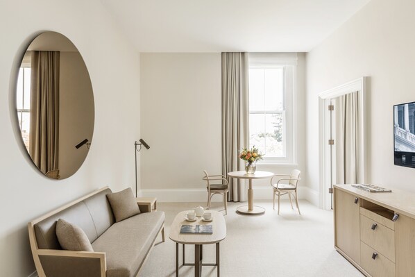 A modern hotel room with a sofa, a small round table with a vase of flowers, a chair, a TV, and a cabinet.