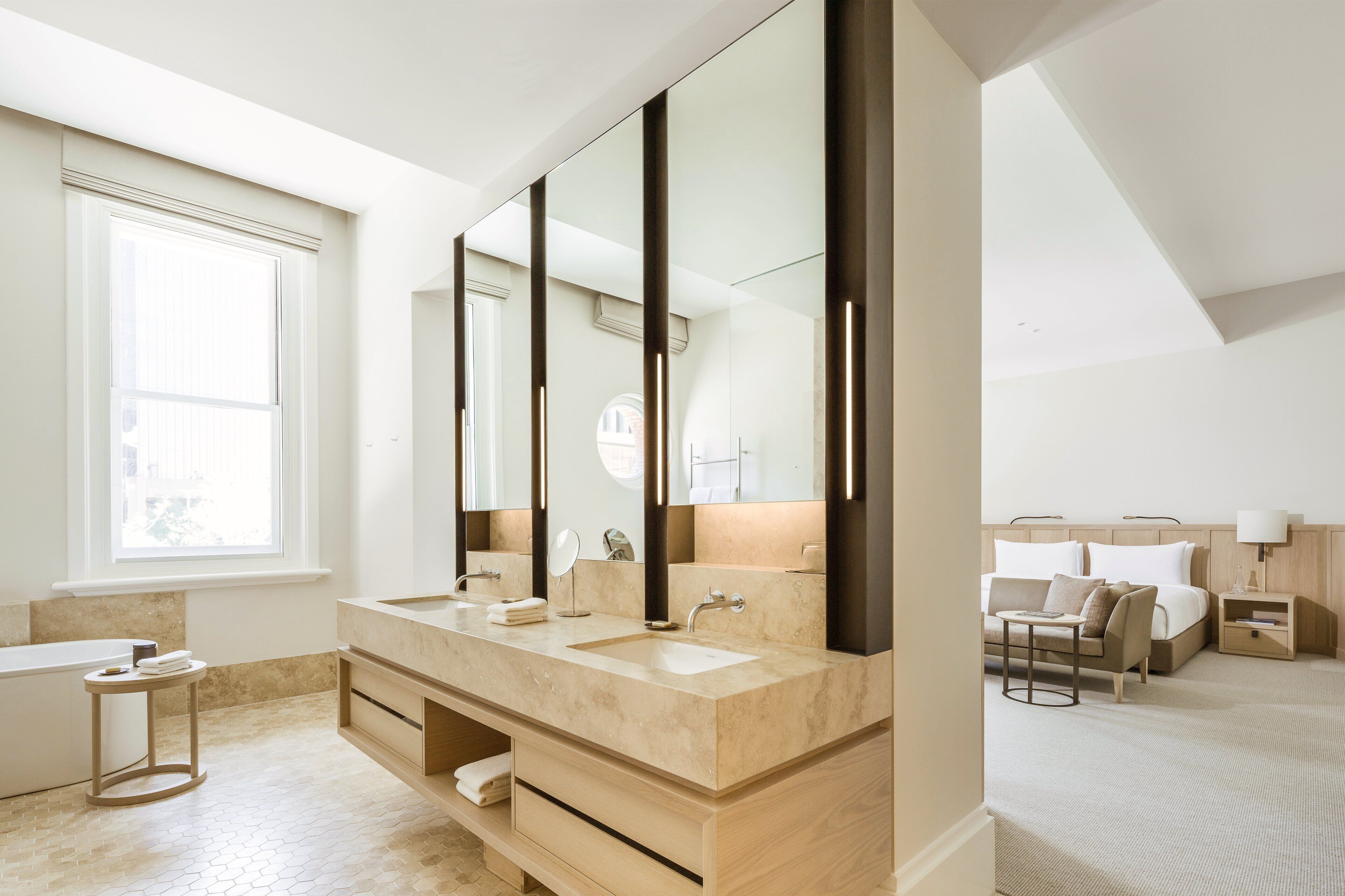 Une chambre d’hôtel moderne dotée d’un grand miroir, de deux vasques, d’une baignoire et d’un lit avec une table de chevet.