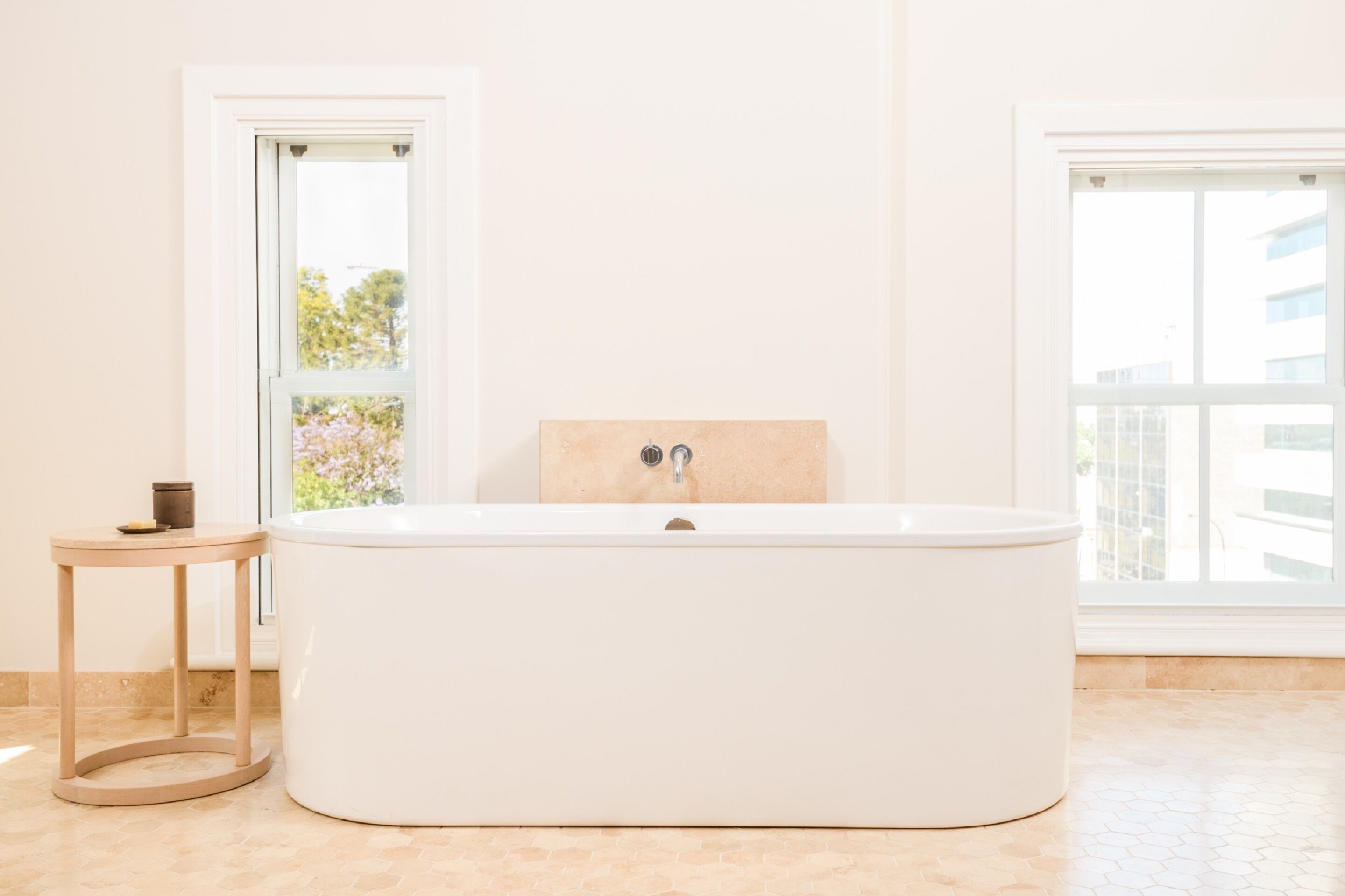 A modern bathroom with a freestanding bathtub, a small round table with a candle, and a window providing a view of trees and buildings.