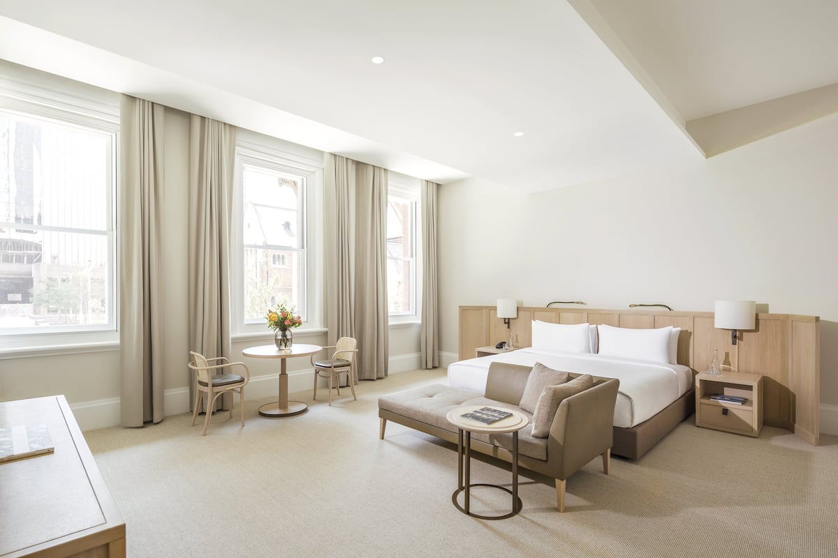 Ein Hotelzimmer mit einem großen Bett, einer Sitzecke mit einer Couch und Sesseln, einem kleinen runden Tisch mit Blumen und einem Schreibtisch mit Stuhl.
