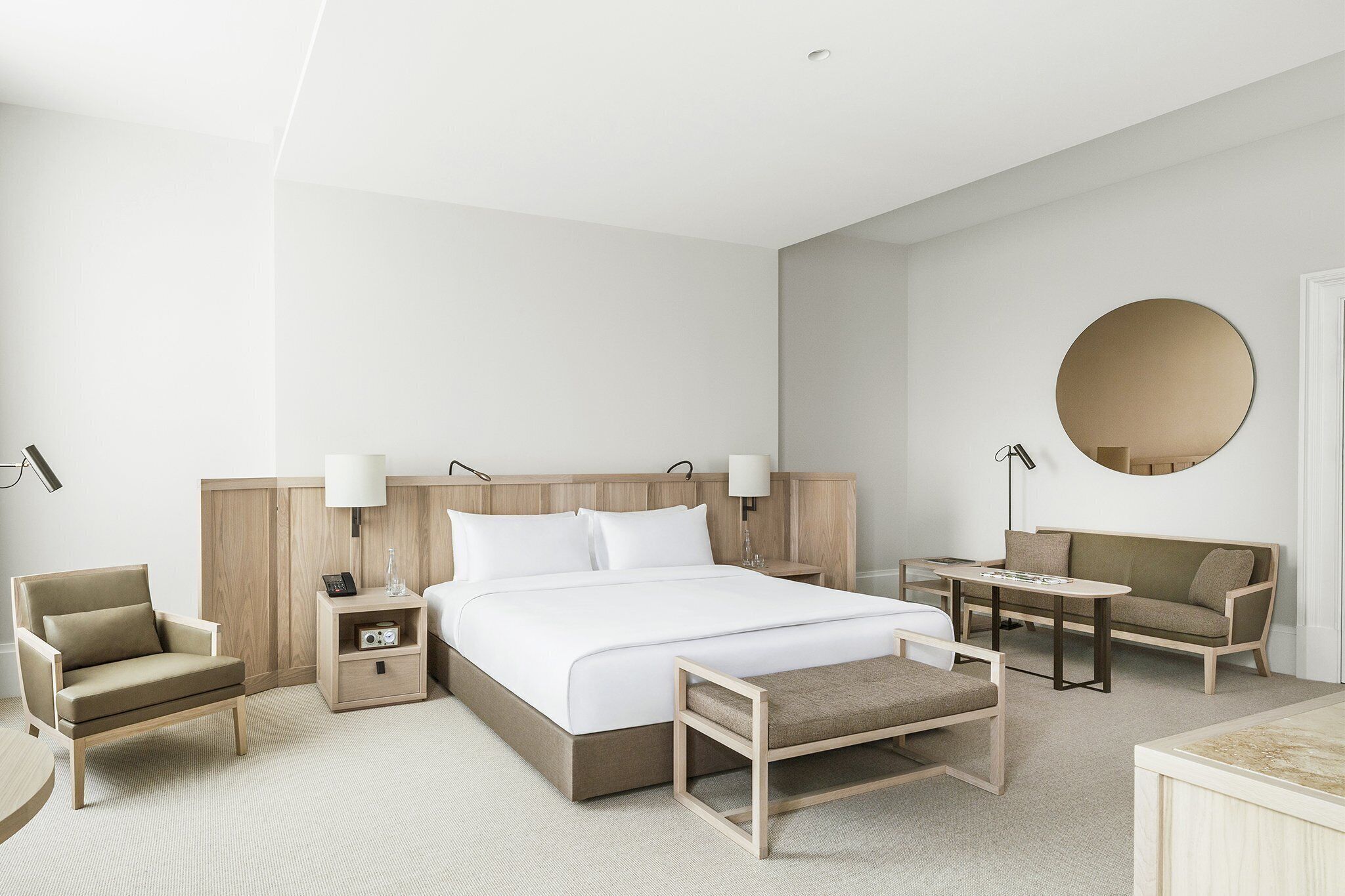 Una habitación de hotel moderna con una cama grande, mesitas de noche, una silla, un sofá y un espejo redondo.