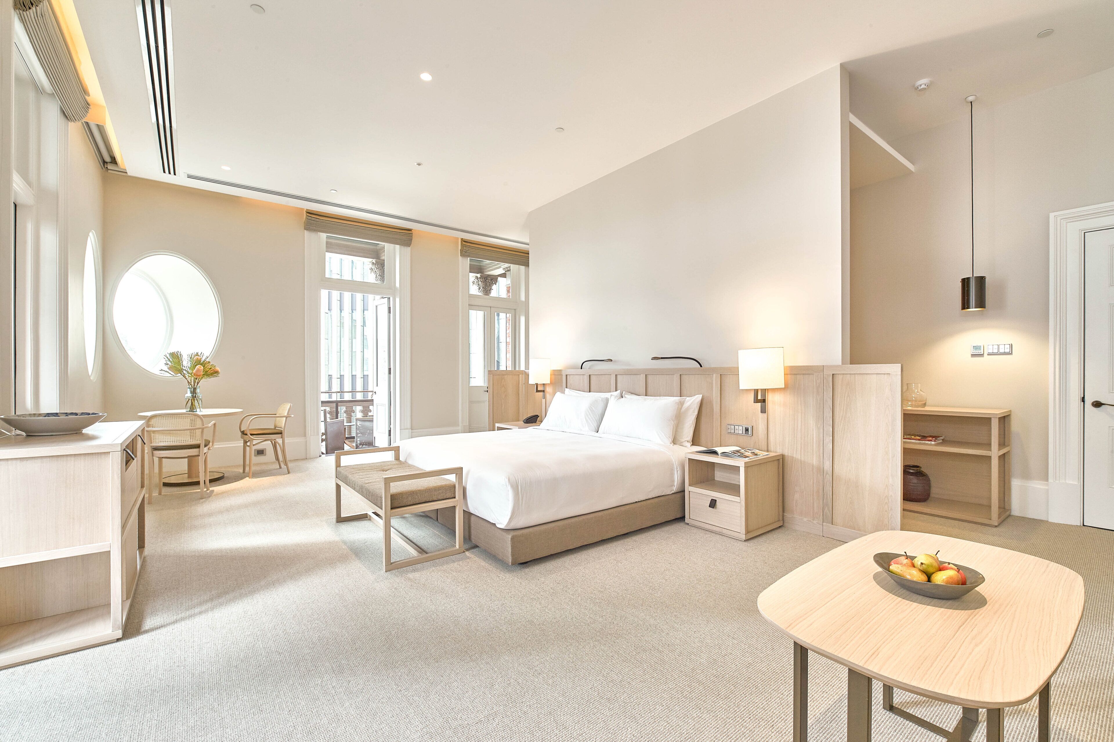 Habitación de hotel con una cama grande, mesitas de noche, un escritorio con una silla, un espejo redondo y una pequeña mesa con fruta.