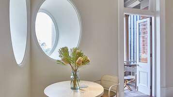 Una mesa redonda con un florero, un espejo redondo y una silla de mimbre en una habitación con una puerta blanca y vistas al exterior.