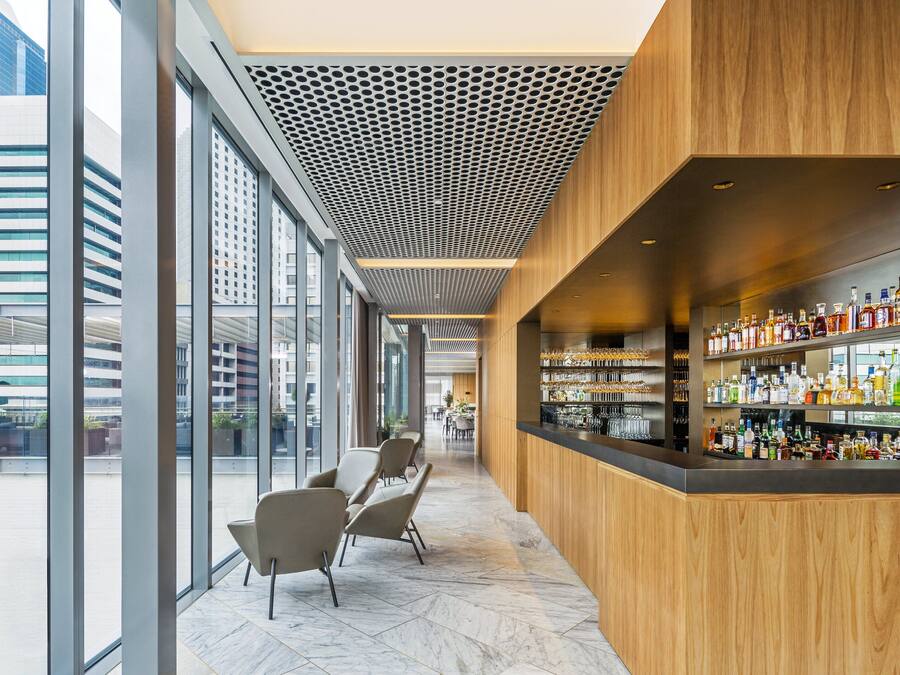 A modern bar area with a wooden counter and shelves of bottles, large windows, and seating.
