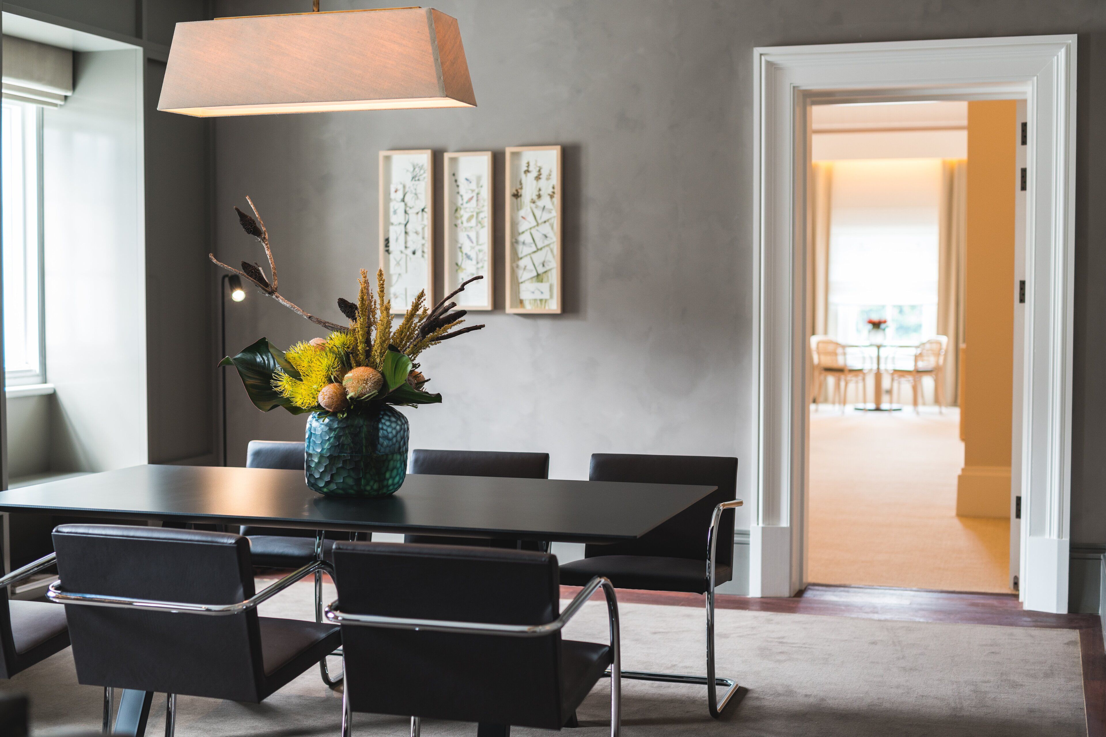 A modern dining area with a table, chairs, and a vase with flowers. There are framed pictures on the wall and a doorway leading to another room.