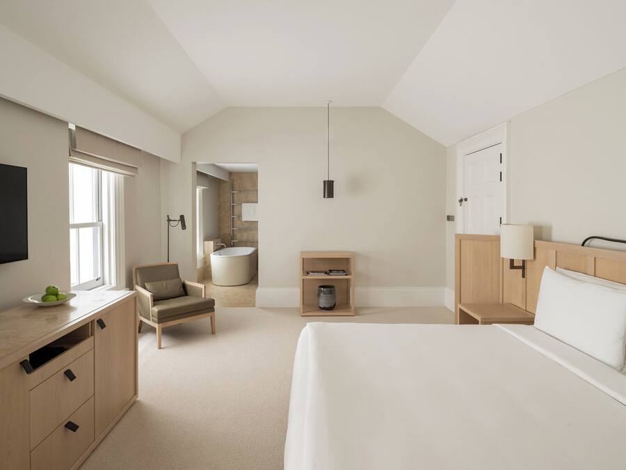 A modern hotel room with a large bed, a desk with a flat-screen TV, a chair, a small table, and a bedside lamp.