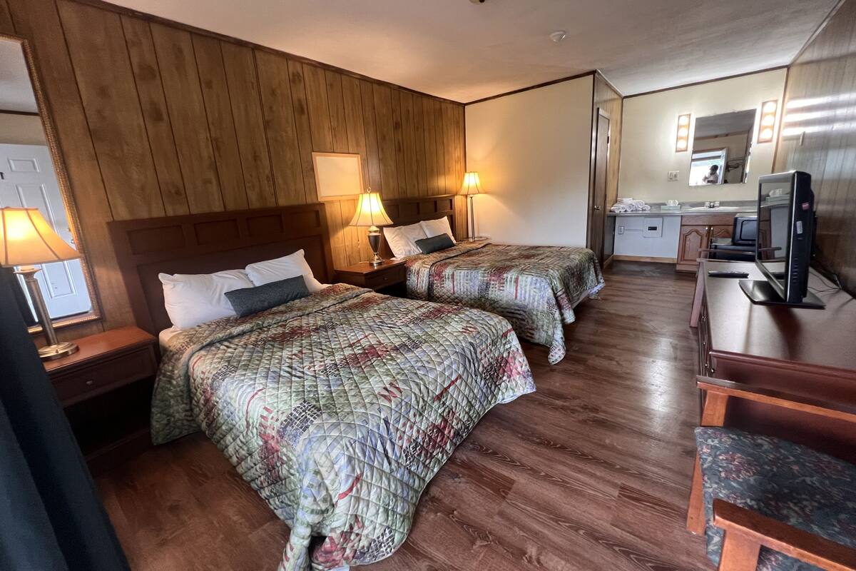 A hotel room with two beds, wooden flooring, a desk with a computer, and a kitchenette in the background.