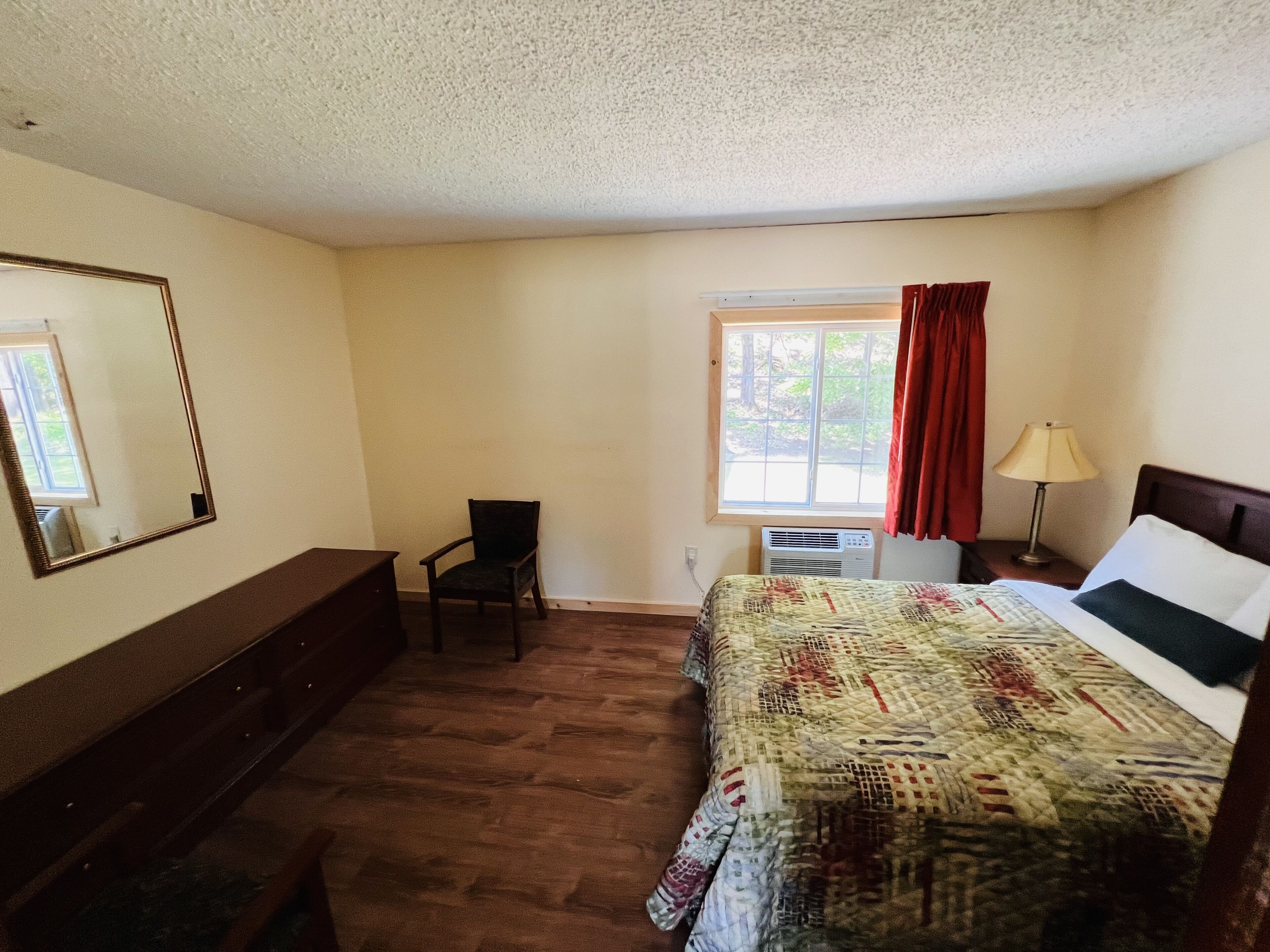 A bedroom with a bed, a chair, a dresser, a window with curtains, and a lamp.