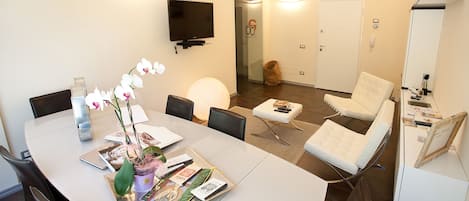 A modern meeting room with a white table, chairs, a TV, and a glass door leading to a hallway.