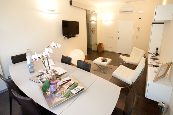 Una sala de reuniones moderna con una mesa blanca, sillas, un televisor y una puerta de vidrio que da a un pasillo.