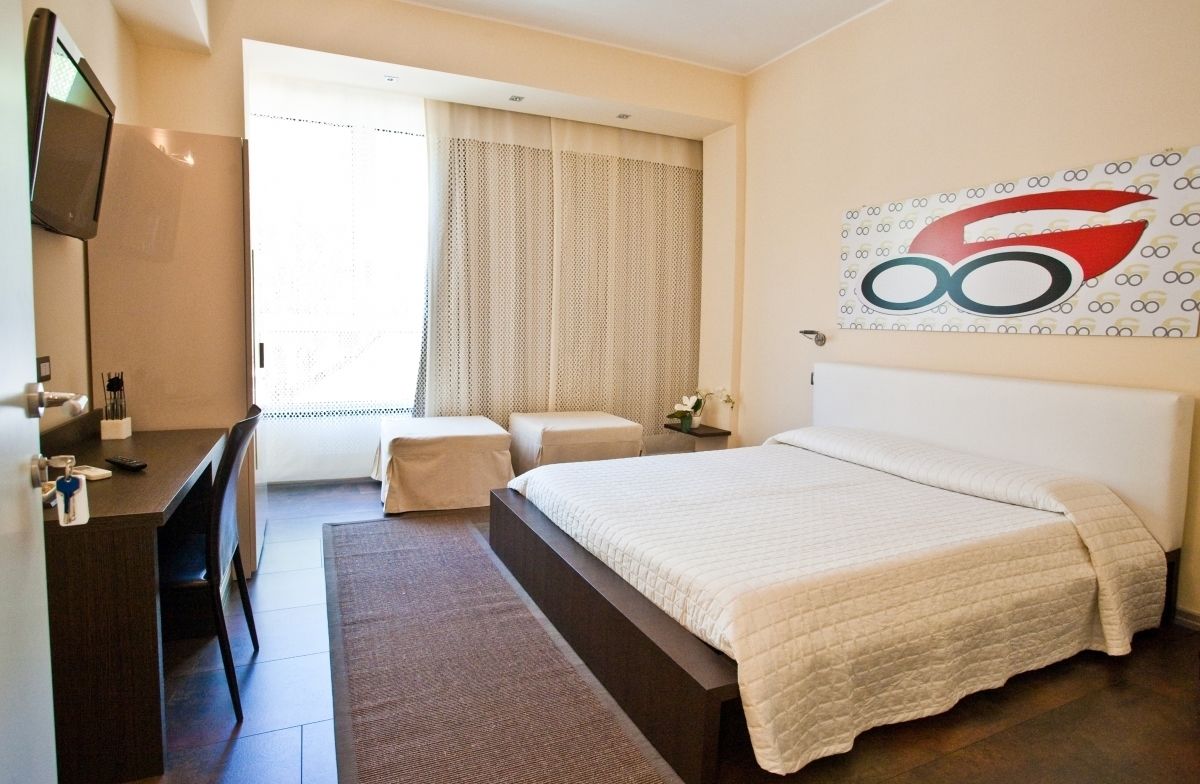 A hotel room with a bed, a desk, a chair, a TV, a window with curtains, and a wall with a logo.