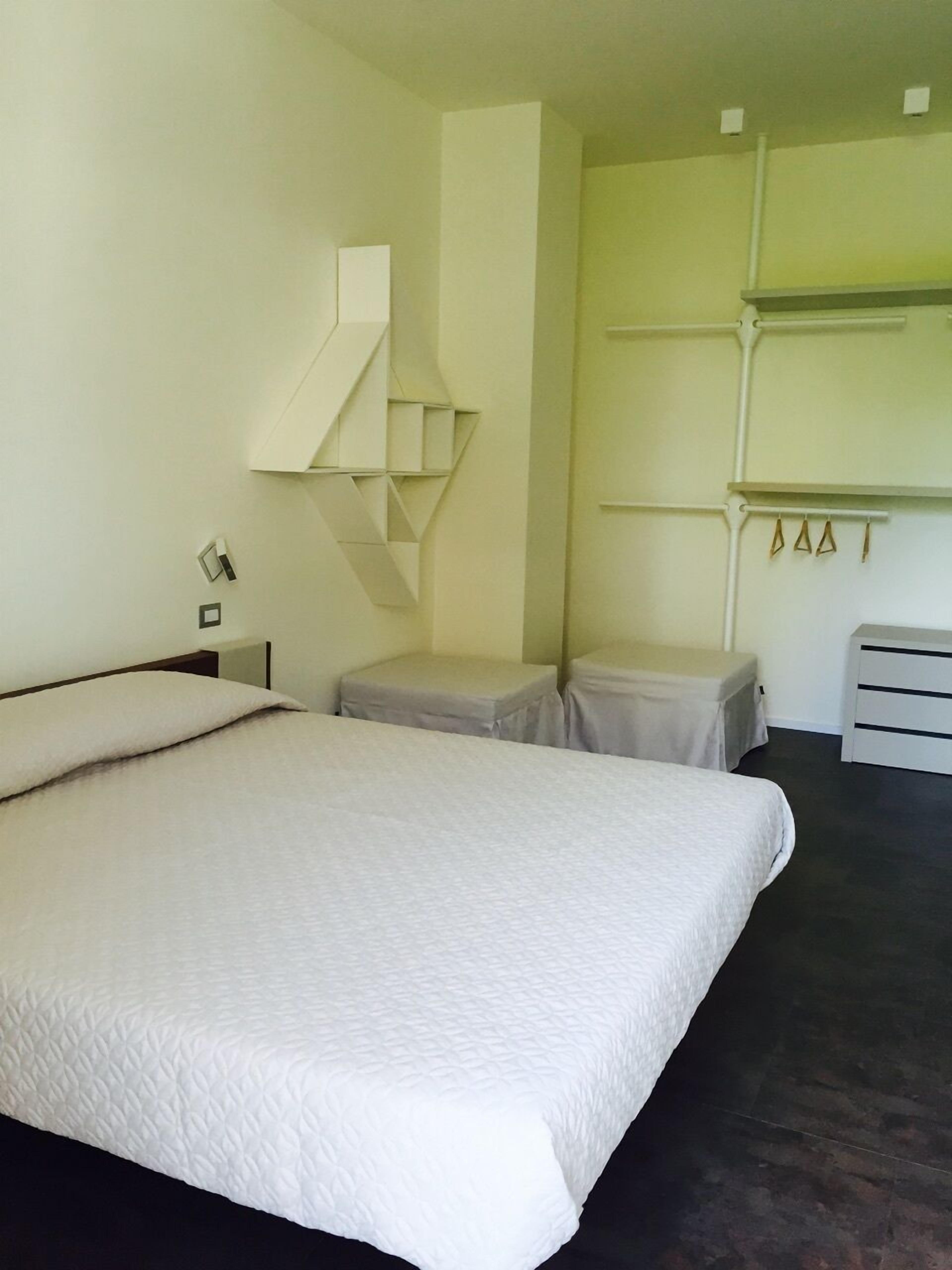 Une chambre simple et moderne, avec un grand lit, deux tables de chevet, un petit tabouret et une étagère intégrée où sont accrochés divers objets.