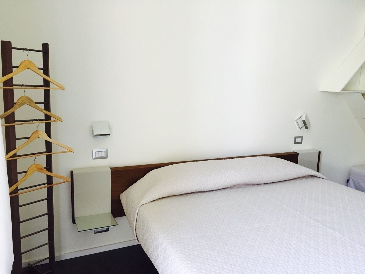 A hotel room with a bed, a wooden hanger rack, a bedside table, and a wall-mounted light.