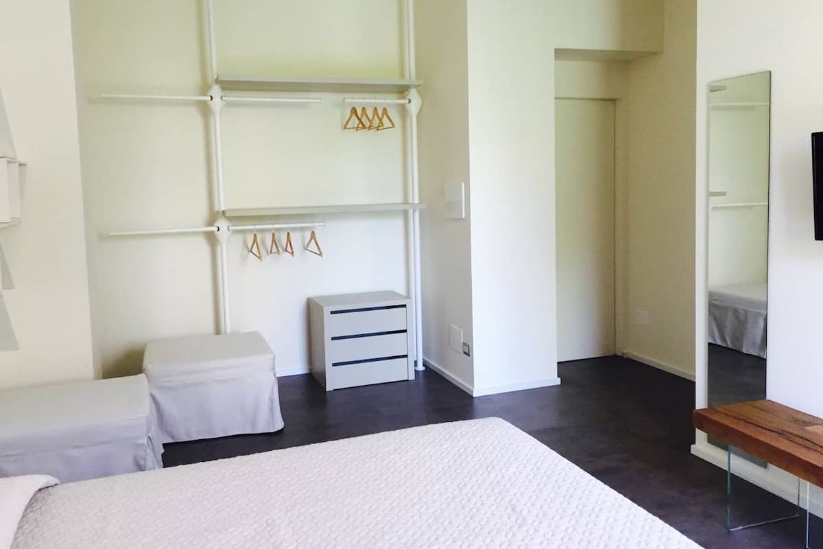 Een slaapkamer met een groot bed, een nachtkastje, een dressoir, een spiegel en een garderobe met daarin hangende kleding.
