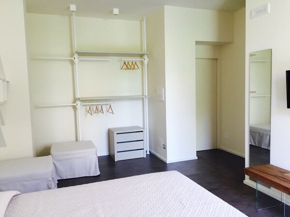 A bedroom with a large bed, a bedside table, a dresser, a mirror, and a wardrobe with hanging clothes. - Cinquecento B&B (Barcellona Pozzo di Gotto)