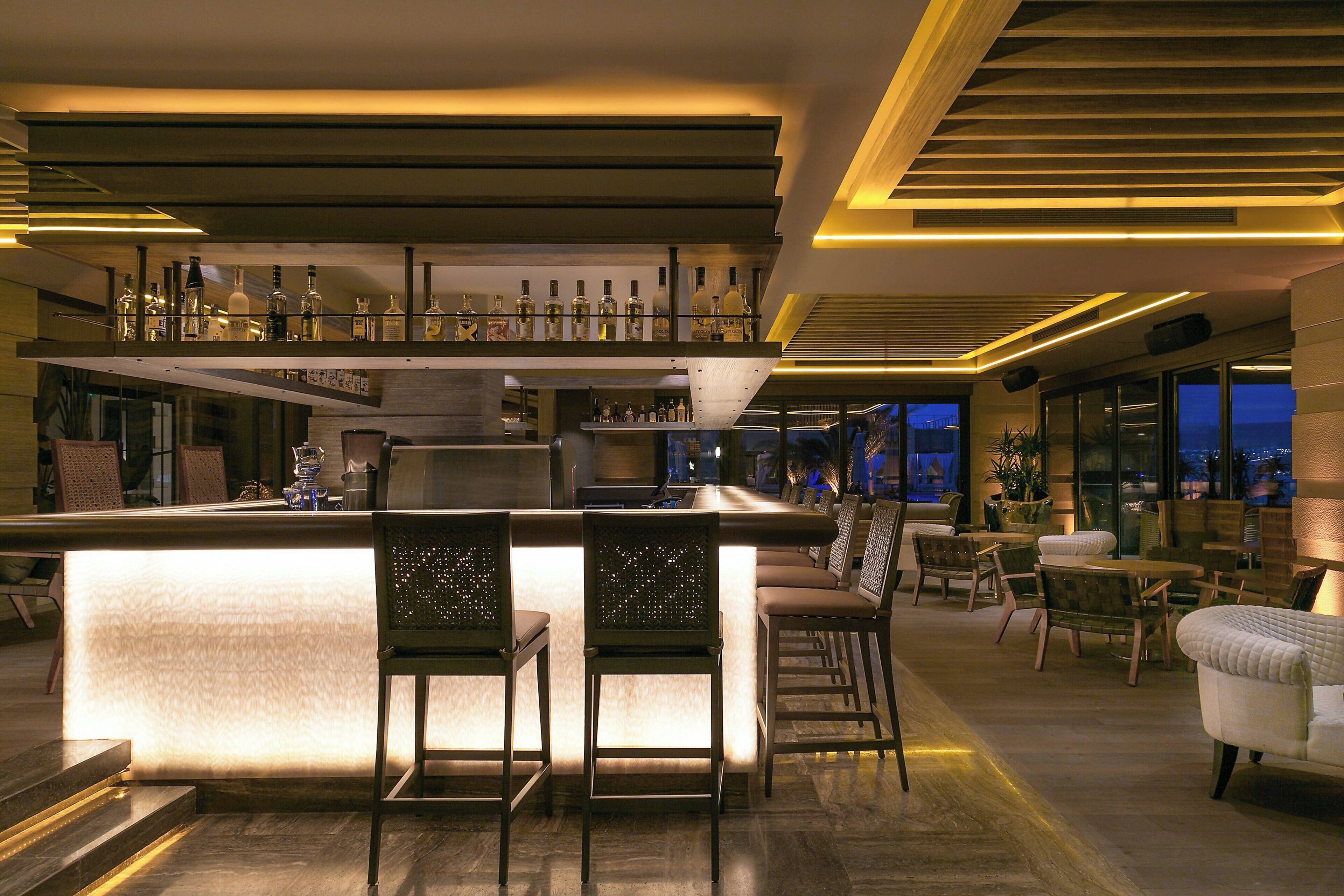 Un espace bar moderne doté d’un comptoir central, de nombreux espaces de détente et d’une vue sur la ville la nuit.