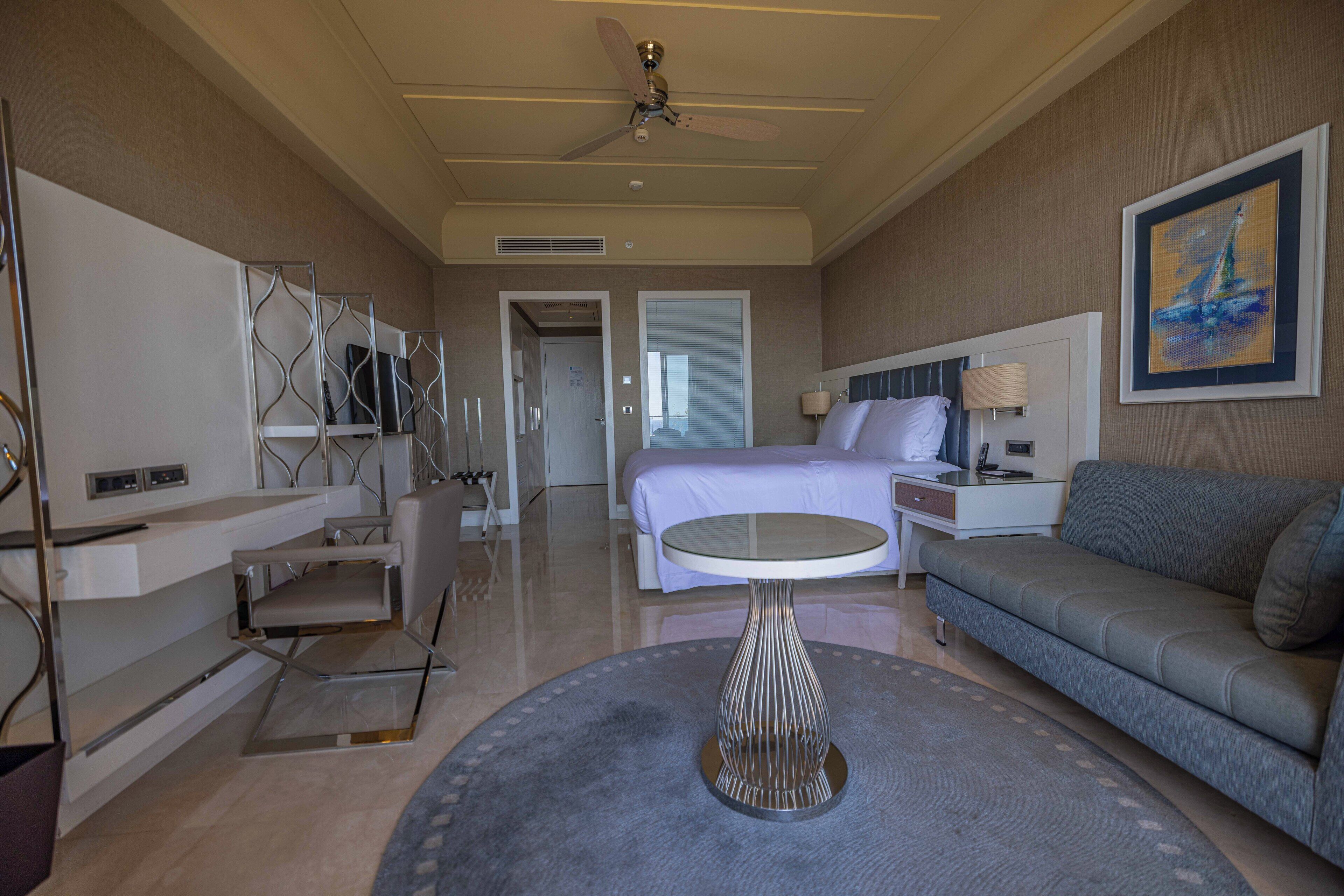 A hotel room with a bed, a sofa, a desk, a chair, a round table, and a ceiling fan.
