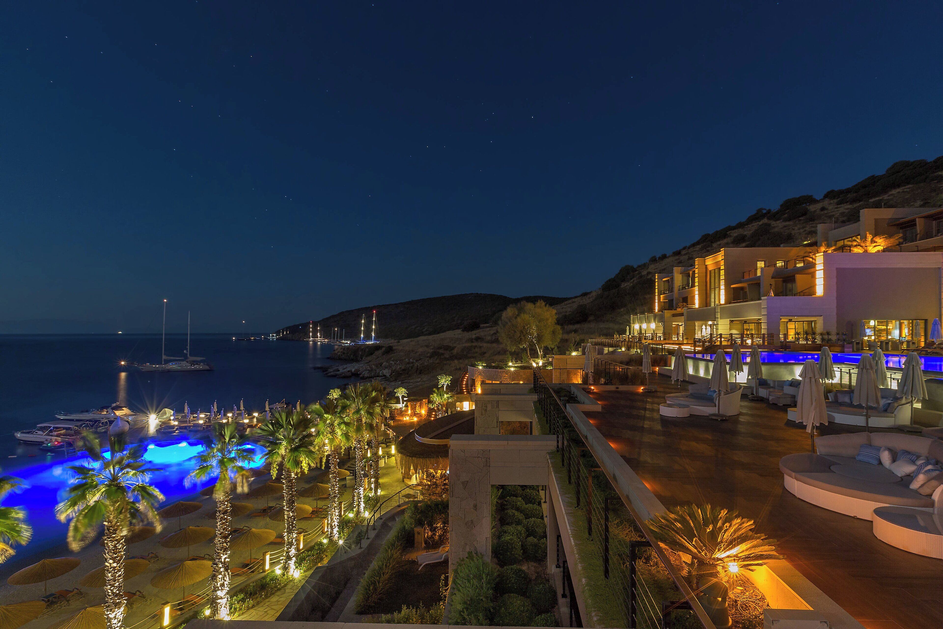 A resort with a pool, palm trees, and a view of the sea at night.