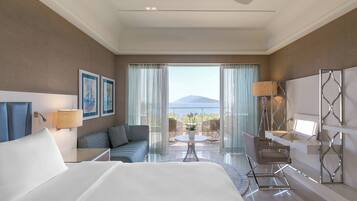 A hotel room with a bed, a sofa, a desk, a chair, a lamp, and a view of the sea.