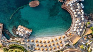 Vista aérea de un complejo turístico costero con un mar turquesa cristalino, una playa de arena y un muelle de madera con sombrillas y tumbonas.