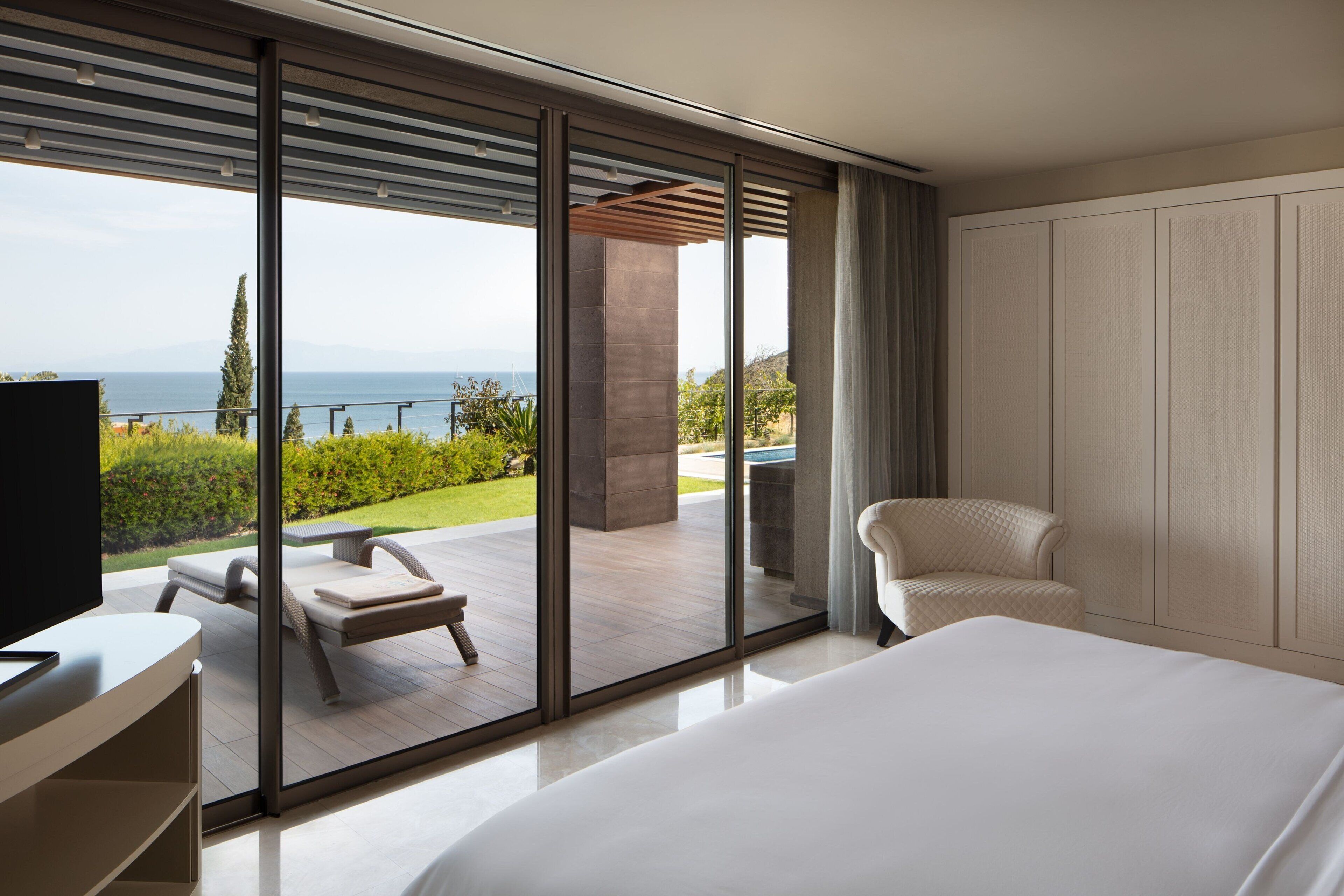 A modern hotel room with a large bed, a TV, and a view of a balcony with outdoor furniture.