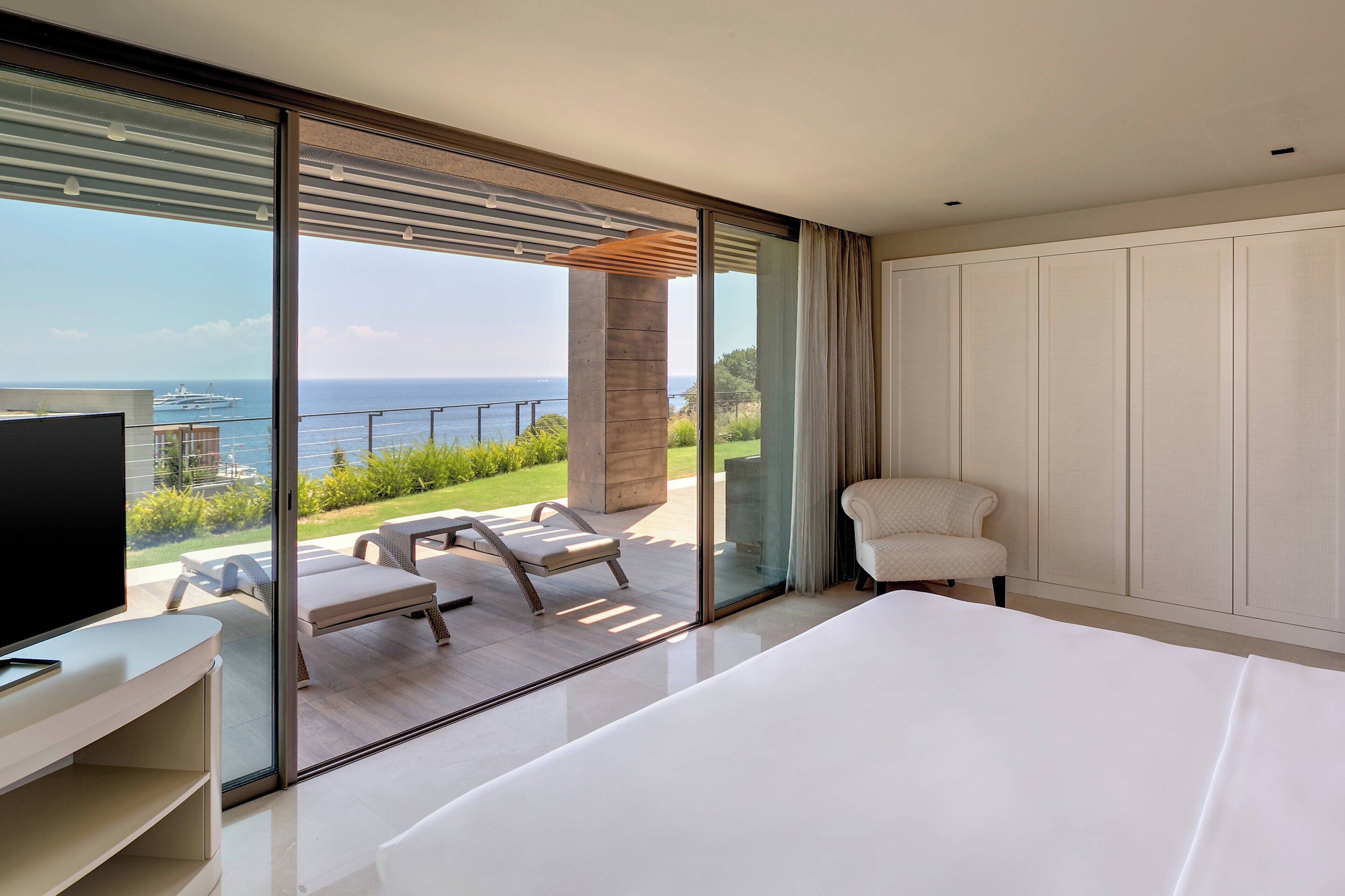 A modern bedroom with a large bed, a TV, and a balcony with a view of the sea.