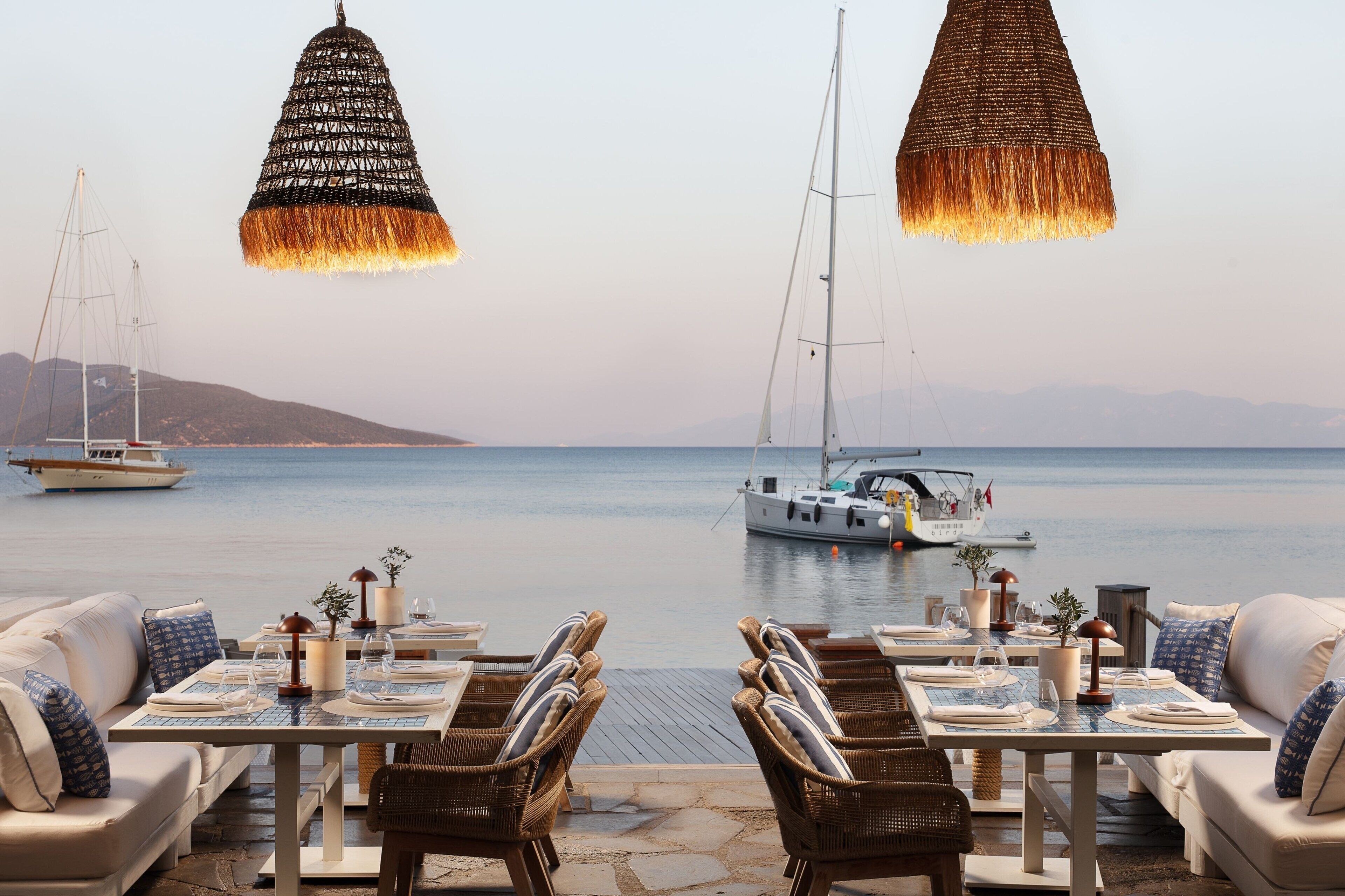 A seaside dining area with tables set for two, overlooking calm waters with boats.