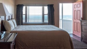 A hotel room with a bed, a desk with a lamp, a dresser, and a view of the sea through a window with curtains. - Copley LaReine Motel (Old Orchard Beach)