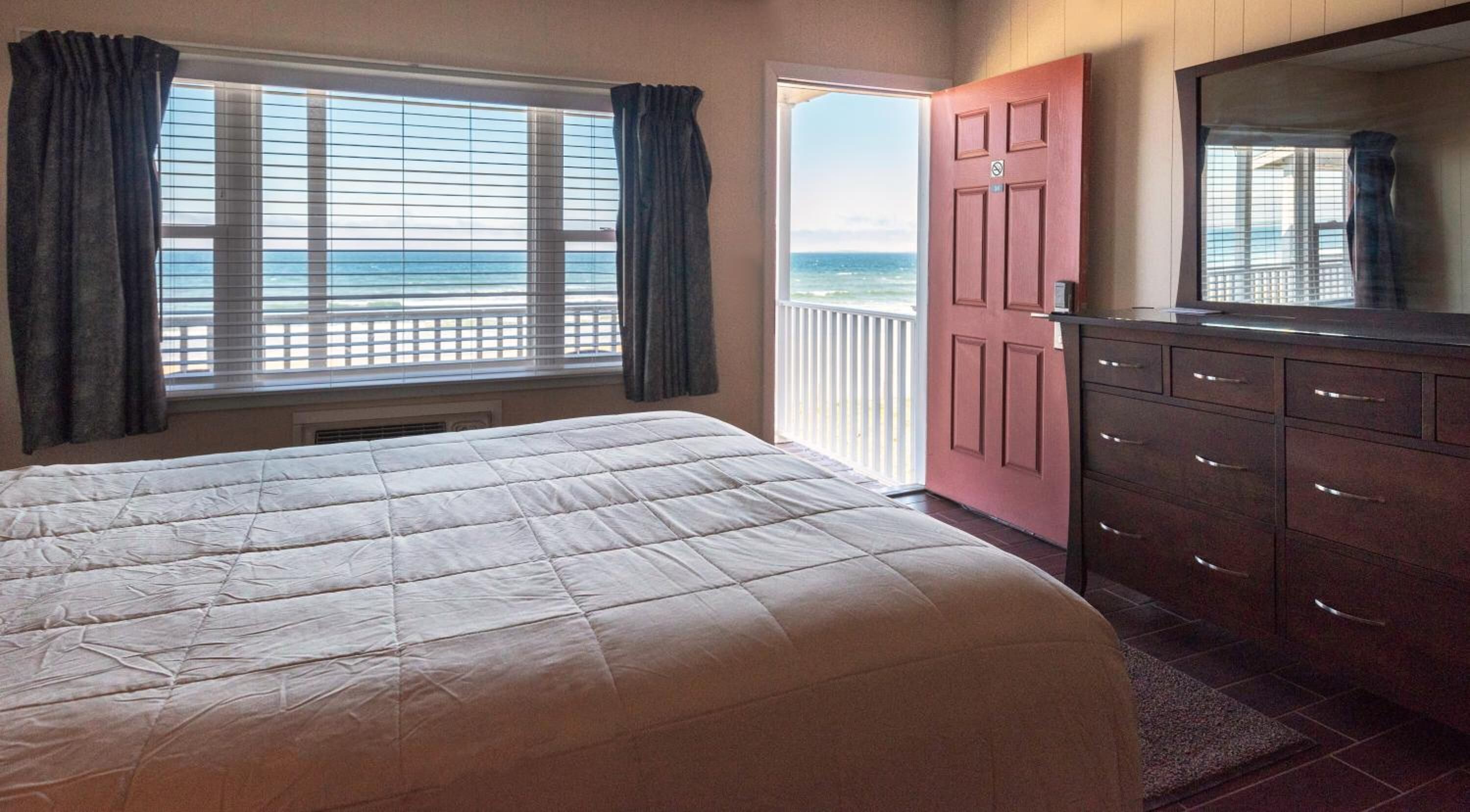 A bedroom with a large bed, a dresser, a mirror, and a door leading to a balcony with a view of the ocean.
