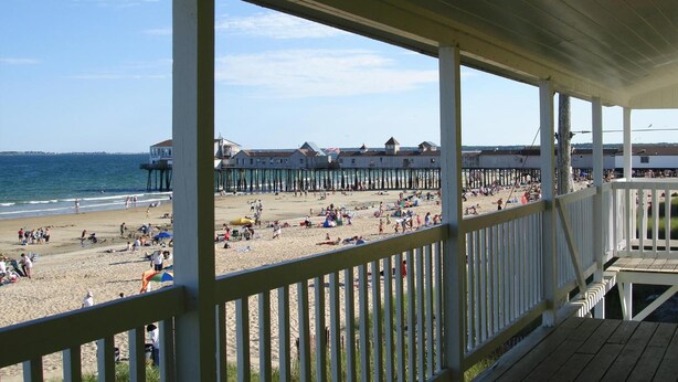 A beach view from a covered wooden deck with a railing, overlooking a sandy beach with people and beach umbrellas. - Copley LaReine Motel (Old Orchard Beach)