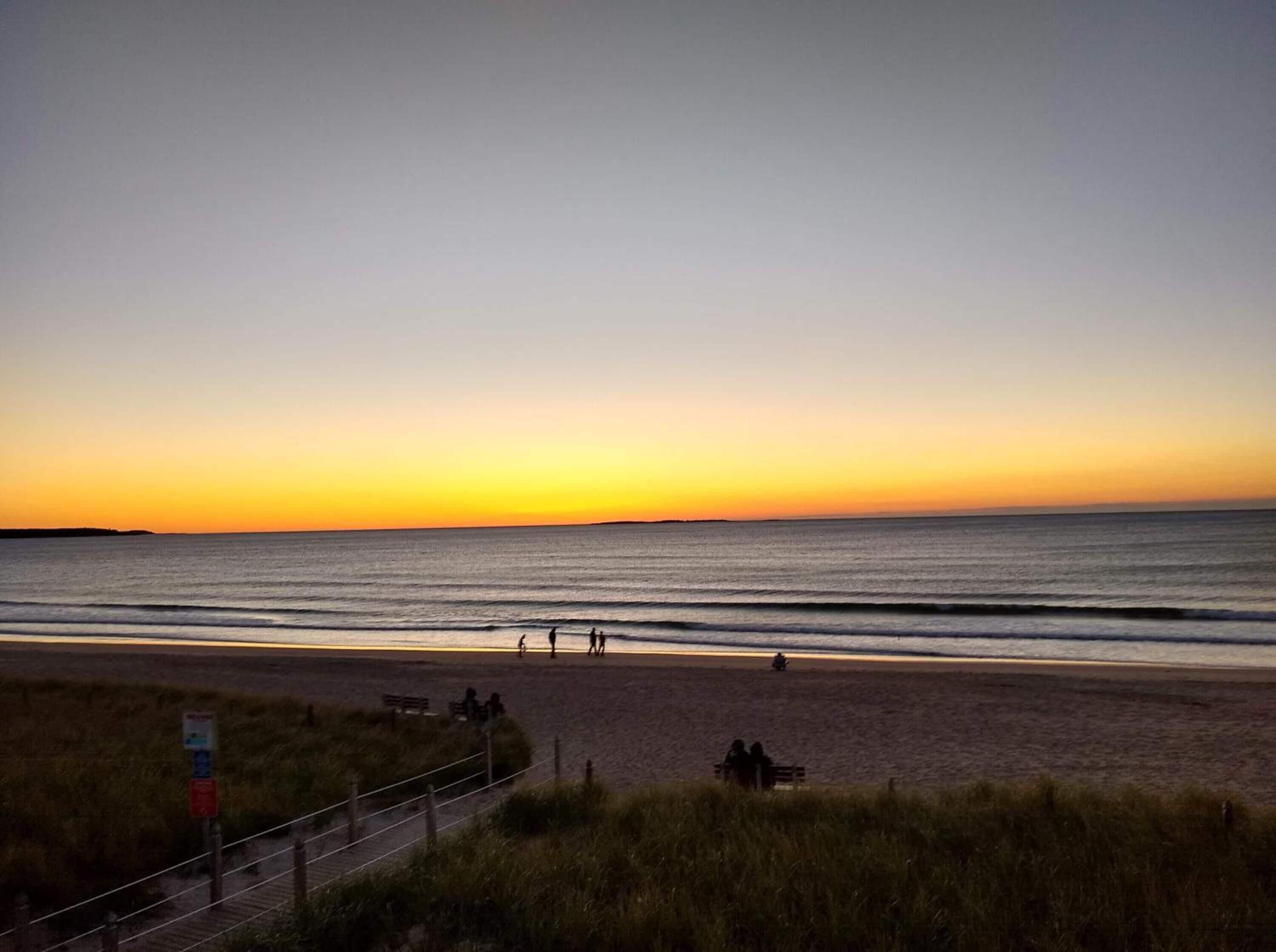 A beach with a sunset, calm sea, and people walking.